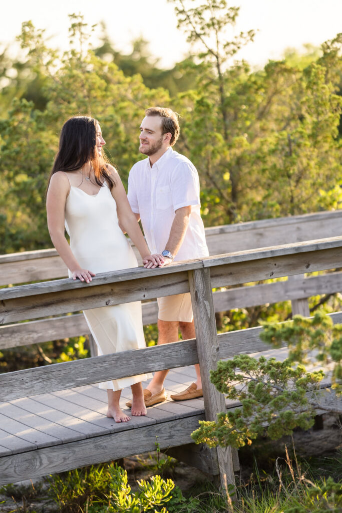 Catie and Zach hold hands and smile at each other on the boardwalk surrounded by coastal greenery at Fire Island National Seashore.