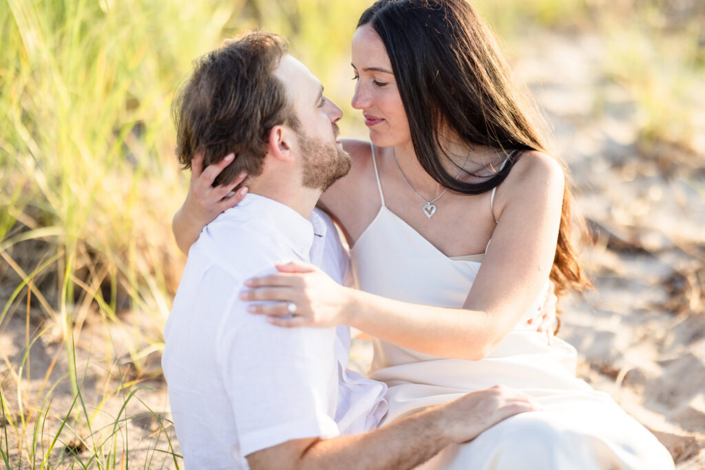Catie and Zach embrace while sitting together in the sand surrounded by beach grass at Fire Island National Seashore.