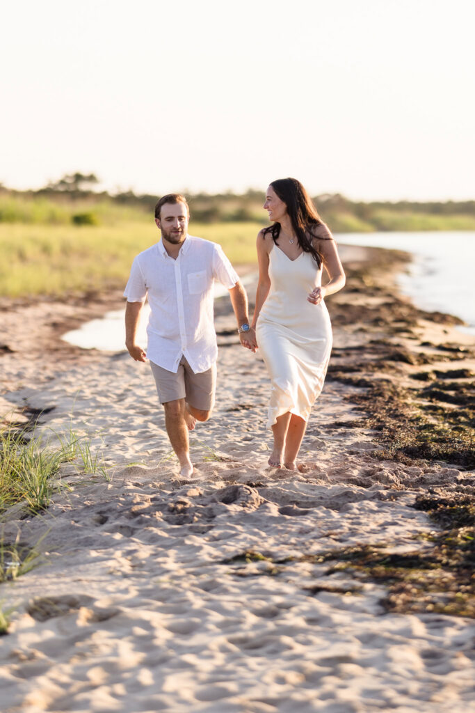 Catie and Zach laugh while walking hand in hand along the sandy shoreline during their Fire Island engagement session.