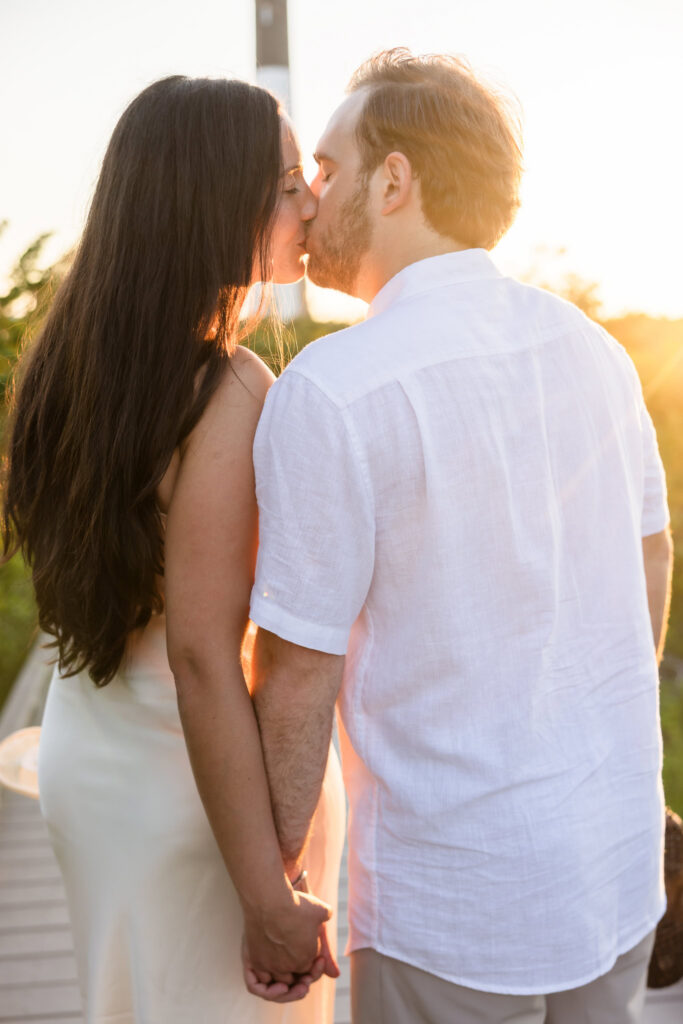 Couple sharing a kiss on the boardwalk near the Fire Island Lighthouse with soft golden backlighting.