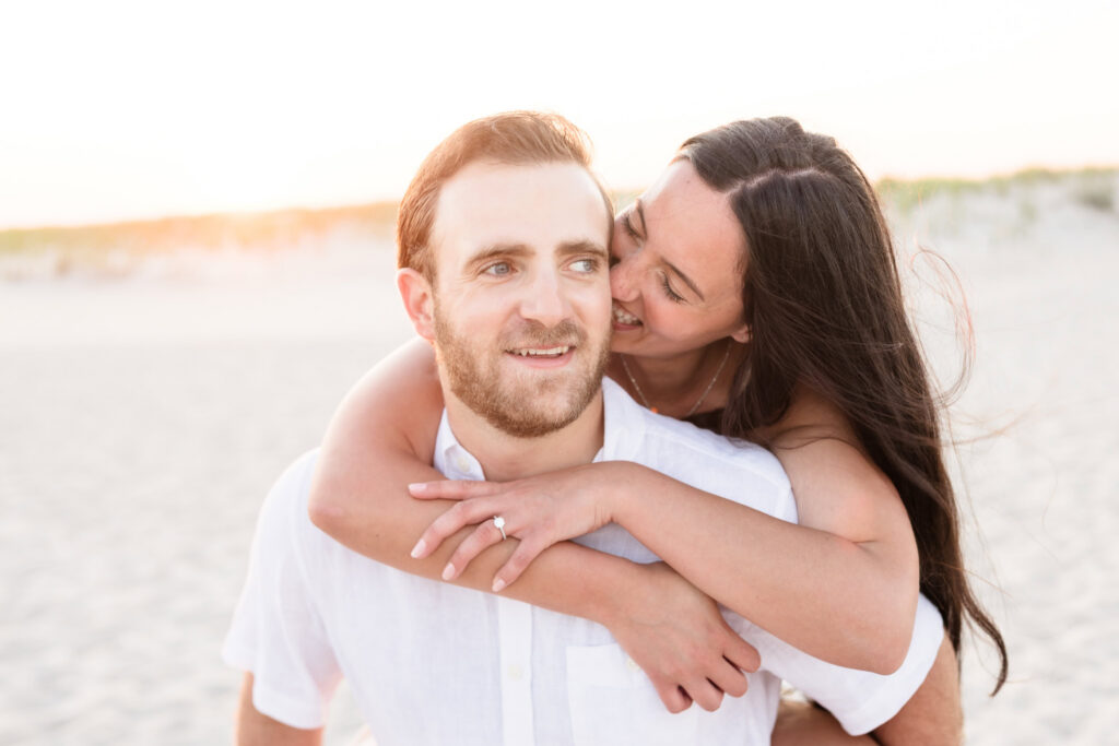 Catie hugs Zach from behind during their Fire Island National Seashore engagement session as golden sunset light glows across the beach dunes.