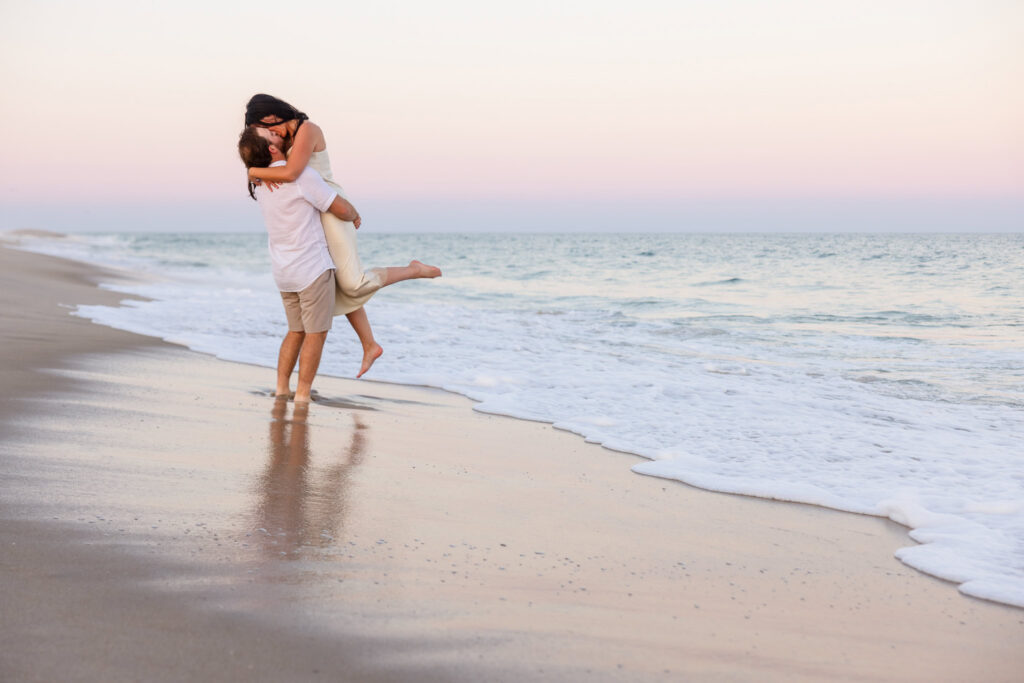 Zach lifts Catie in the surf during their sunset engagement session at Fire Island National Seashore with pastel skies overhead.