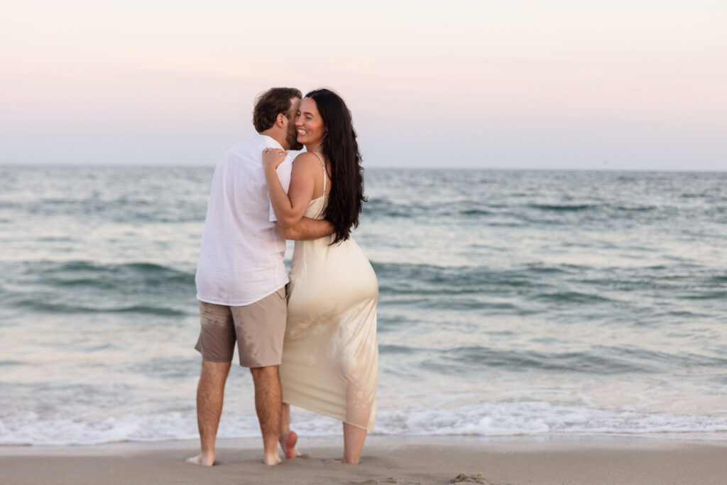 Catie smiles while embracing Zach on the shoreline during their relaxed Fire Island National Seashore engagement session.