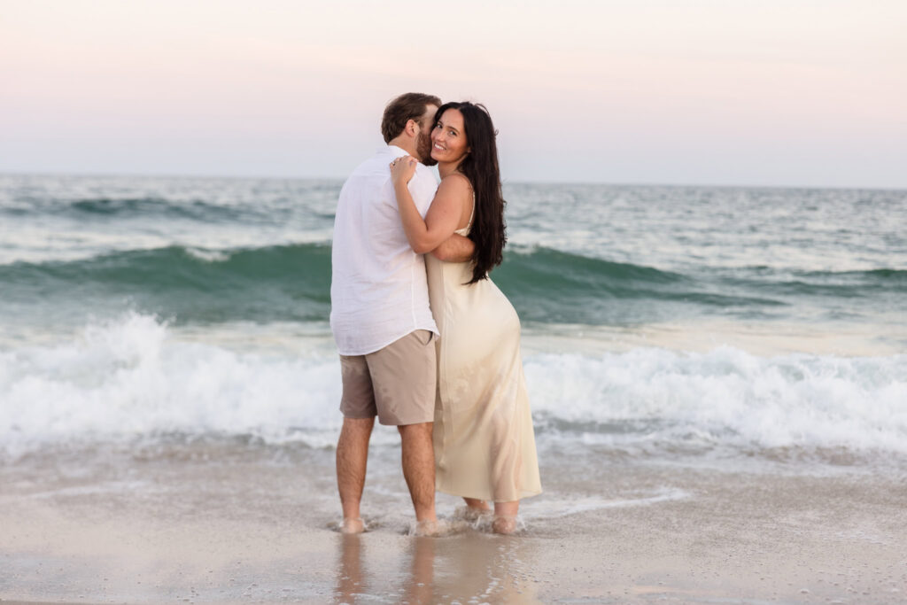 Catie smiles toward the camera as she hugs Zach with rolling ocean waves behind them at Fire Island National Seashore.