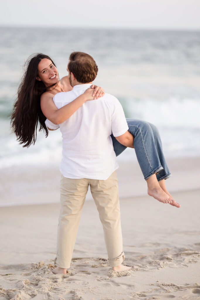 Zach carries Catie along the beach during their engagement session as she smiles toward the camera.