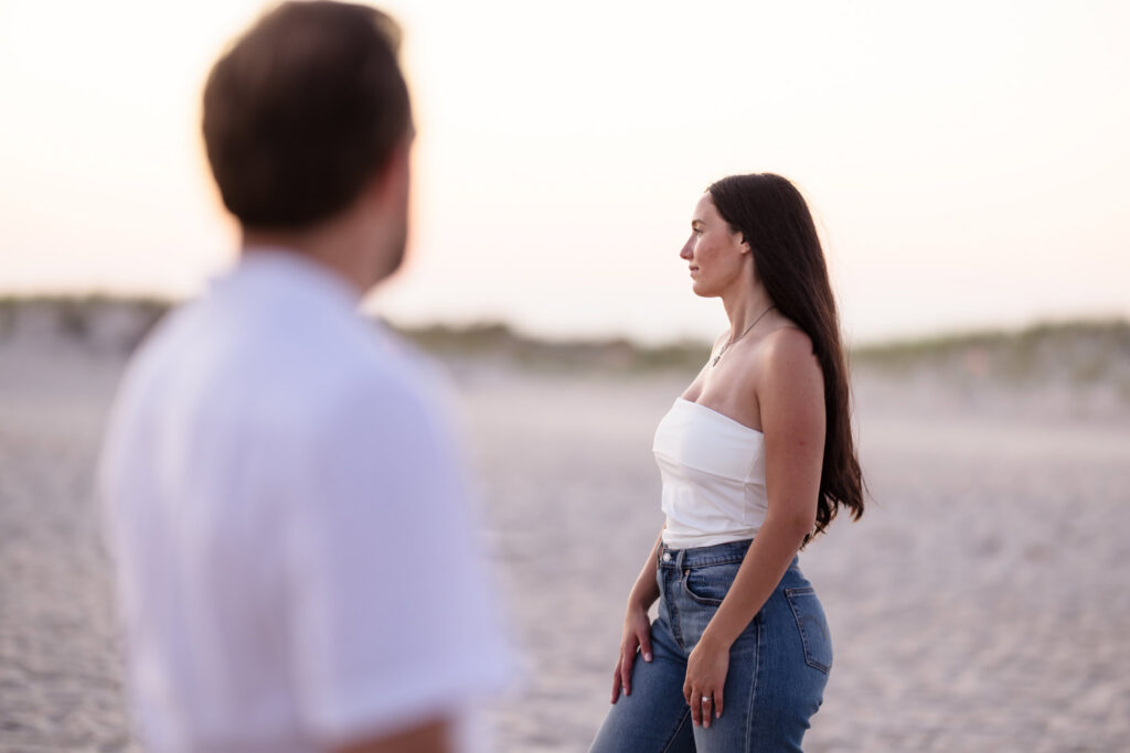 Catie stands quietly on the beach as Zach looks toward her during their peaceful engagement session.