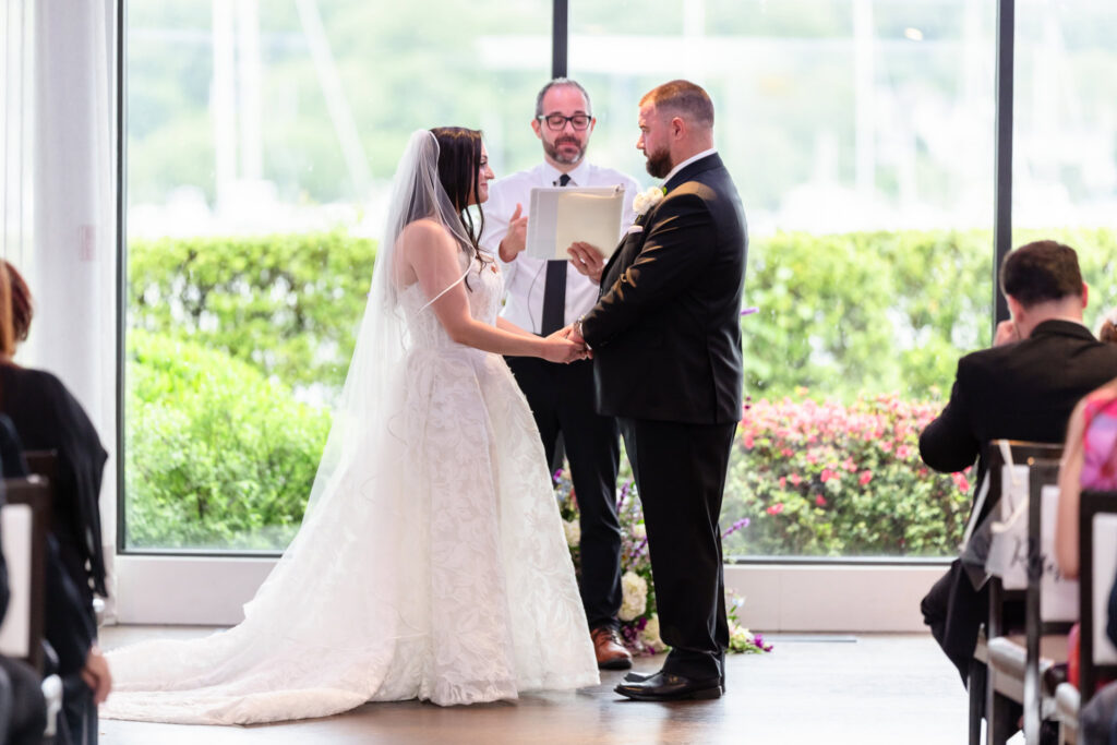Bride and groom exchanging vows during their indoor Harbor Club at Prime wedding overlooking Huntington Harbor.