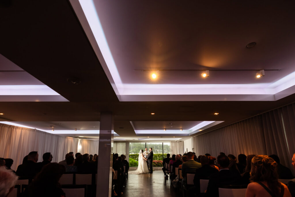 Indoor ceremony during a Harbor Club at Prime wedding overlooking Huntington Harbor through floor-to-ceiling windows as the couple exchanges vows.