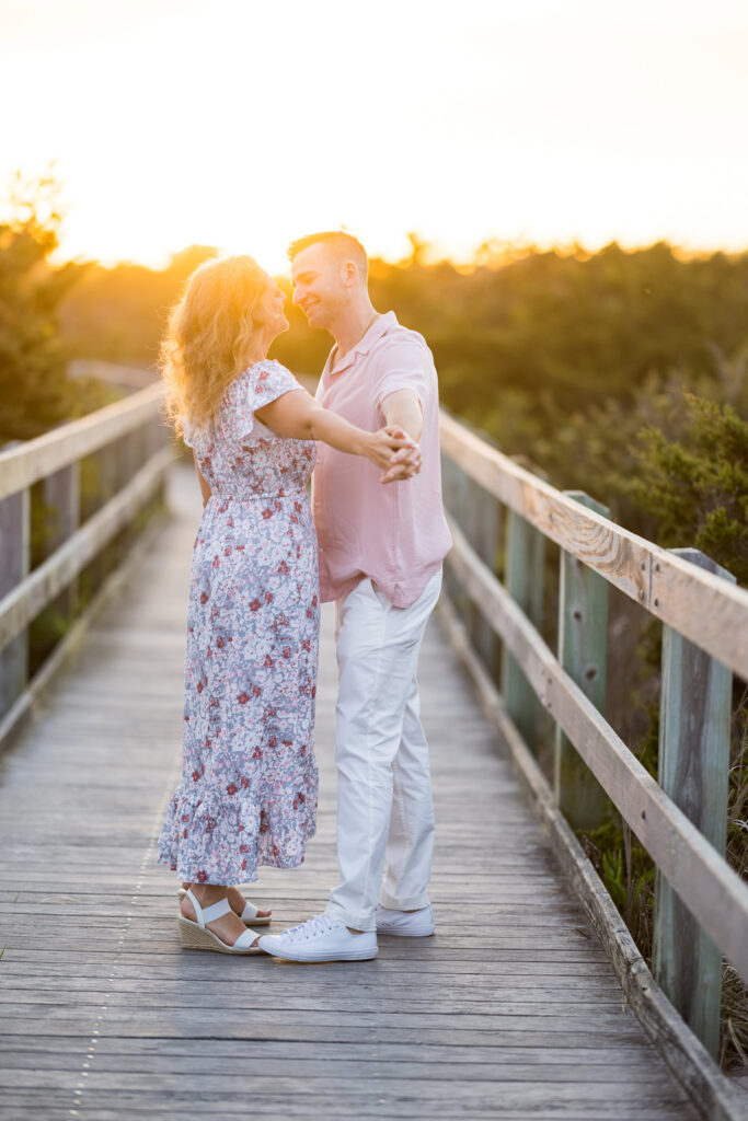 Couple holding hands and smiling on the Fire Island boardwalk in warm golden hour light.