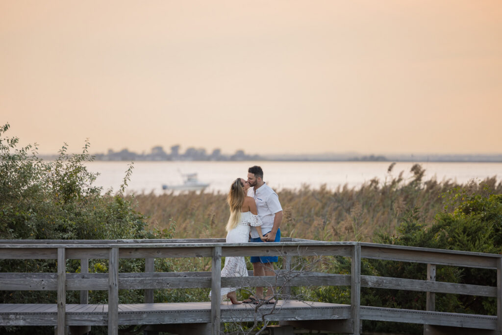 Couple sharing a kiss on a wooden boardwalk overlooking the bay near Fire Island National Seashore or Robert Moses State Park engagement photos