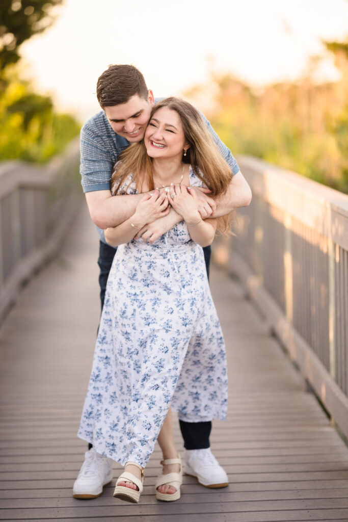 Couple embracing playfully on the boardwalk with soft evening light and dune vegetation behind them.