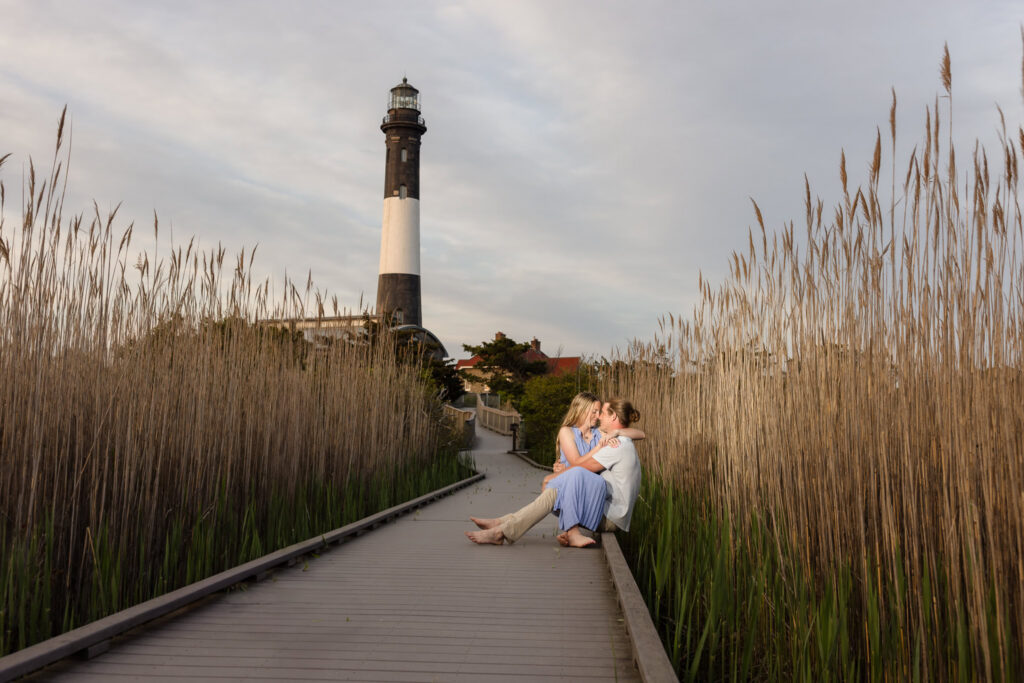Couple seated together on the boardwalk with the Fire Island Lighthouse rising behind them.