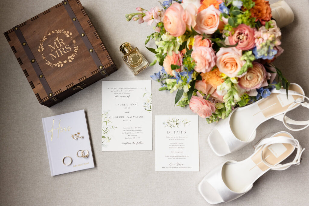 Wedding invitation suite, bridal shoes, bouquet, perfume, rings, and keepsake box arranged for detail photographs before Lauren and Joe's Watermill Caterers wedding.