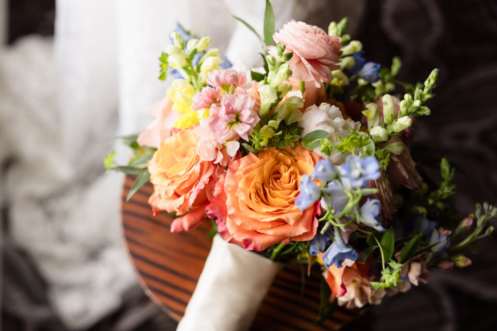 Colorful bridal bouquet featuring peach roses, ranunculus, blue delphinium, and soft pink flowers before the wedding day.