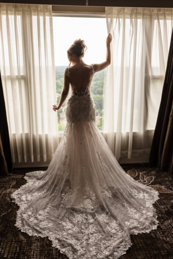 Lauren stands in front of a hotel window in her lace wedding dress before her Watermill Caterers wedding.
