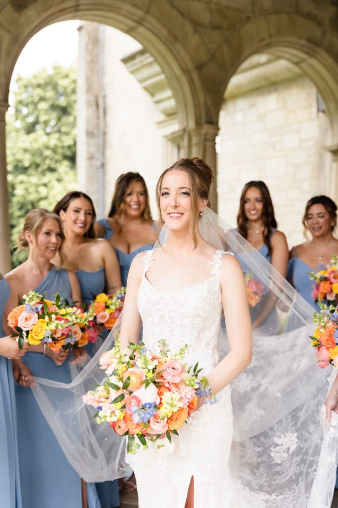 Bridal portrait of Lauren holding her bouquet with bridesmaids gathered behind her under the stone arches at Planting Fields Arboretum.