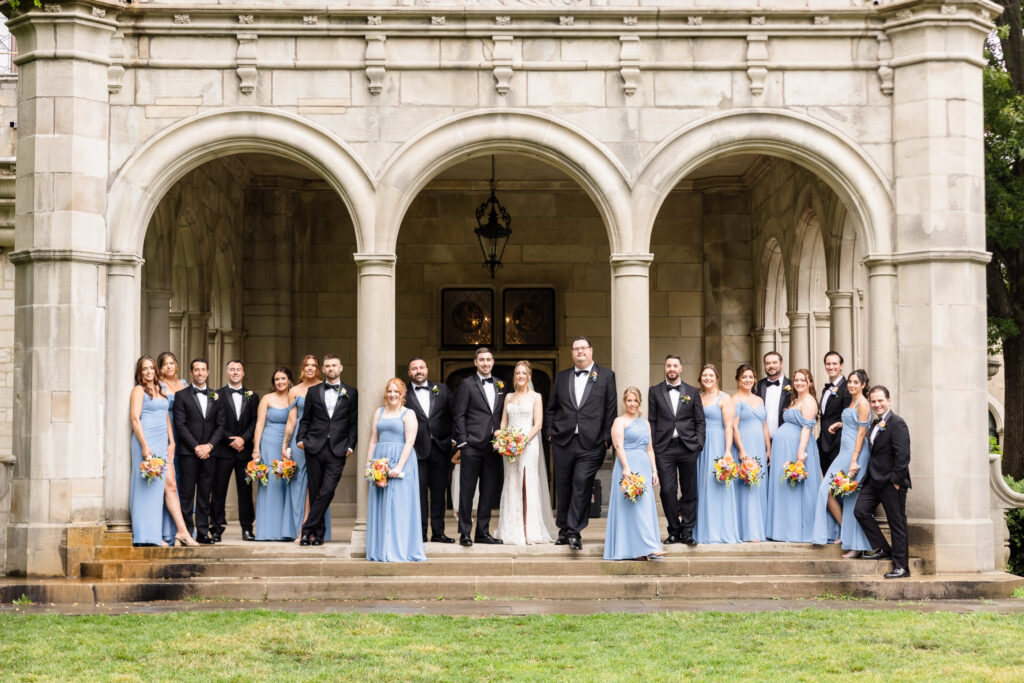 Full wedding party gathered on the grand stone entrance steps of Planting Fields Arboretum on a rainy June wedding day.