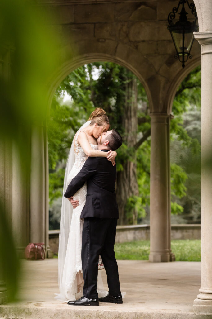 Groom lifting the bride for a kiss beneath the covered stone pavilion at Planting Fields Arboretum during wedding portraits.