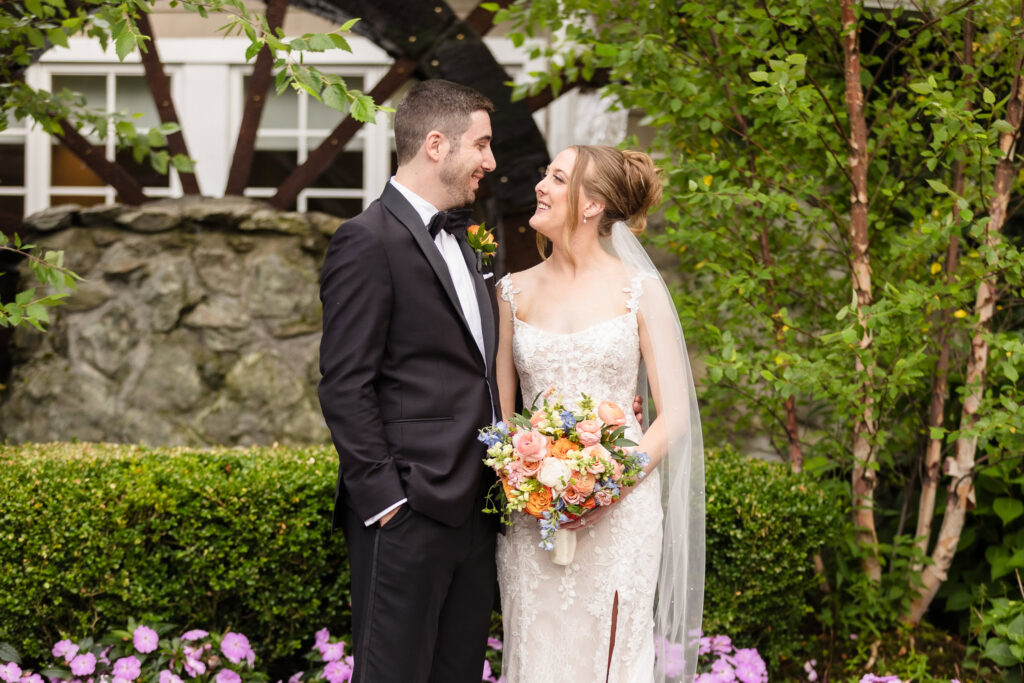 Lauren and Joe smiling at one another during garden portraits surrounded by lush greenery and summer flowers at Planting Fields Arboretum.