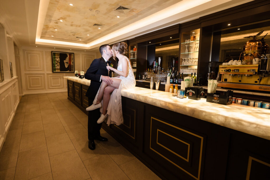Bride and groom sharing a kiss at the illuminated bar inside Watermill Caterers during evening portraits.