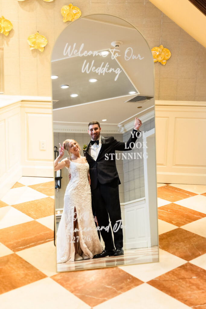 Lauren and Joe posing together in front of their personalized wedding welcome mirror at Watermill Caterers.