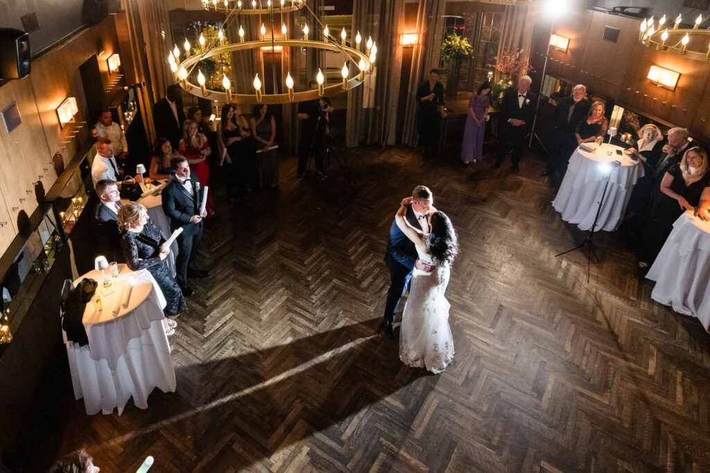 Newlyweds sharing their first dance beneath warm chandelier light as wedding guests gather around them.