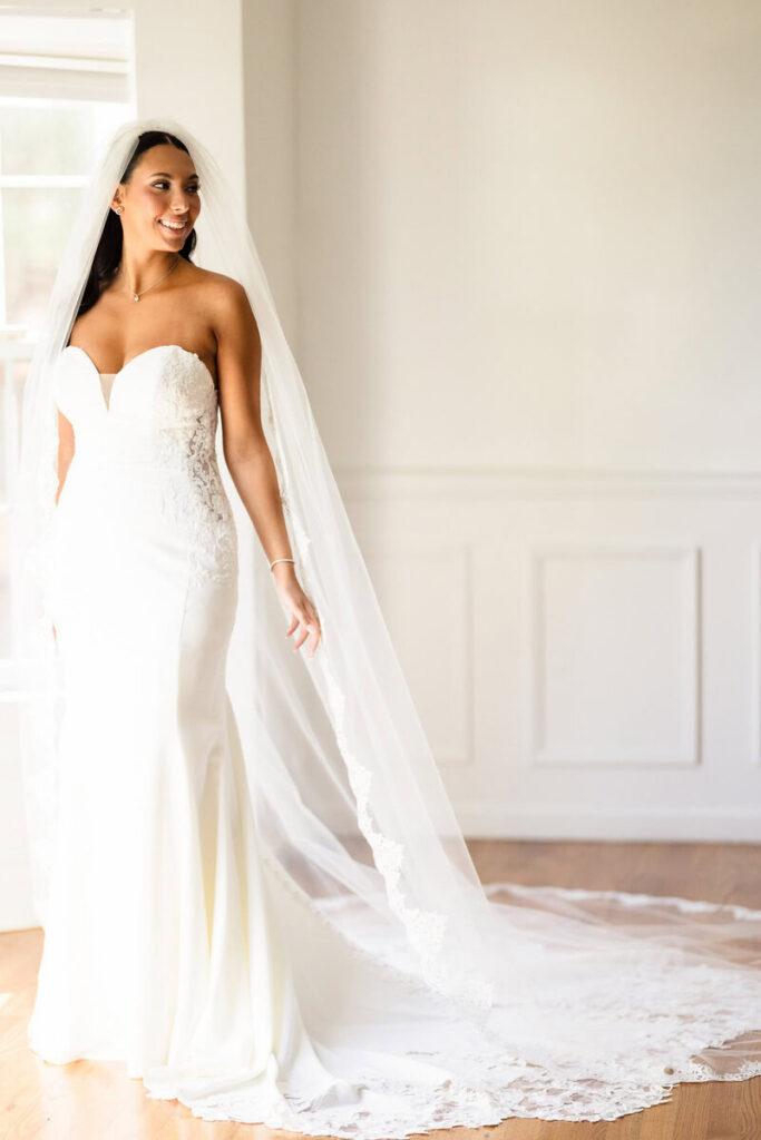 Bride in a lace wedding gown and long veil standing in soft natural window light before her ceremony.