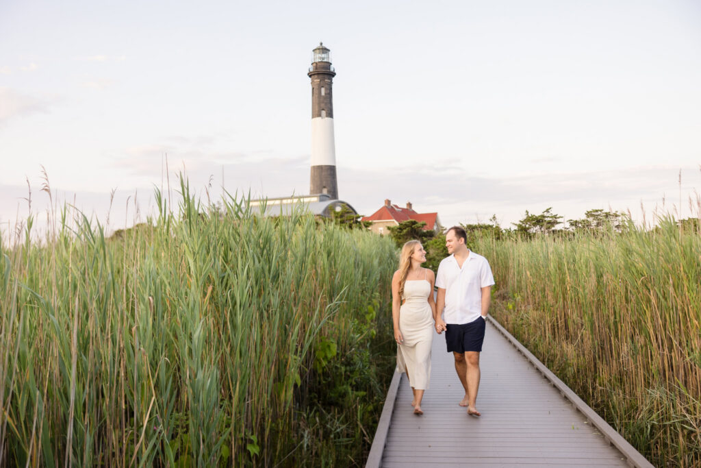 Couple walking hand in hand through tall dune grass with the Fire Island Lighthouse behind them.