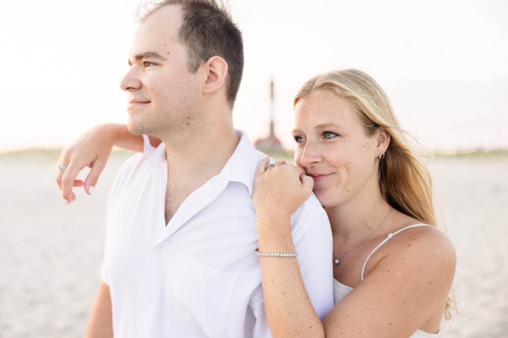 Close portrait of an engaged couple on the beach with the Fire Island Lighthouse softly visible in the distance.