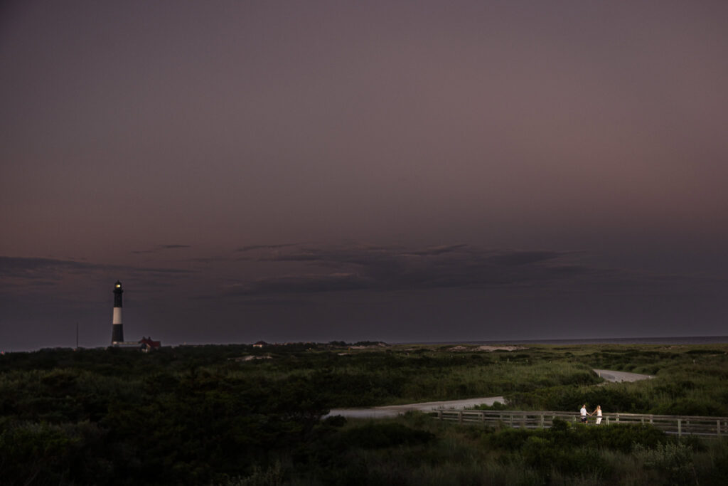 Wide evening view of an engaged couple walking along the boardwalk with the illuminated Fire Island Lighthouse in the distance.