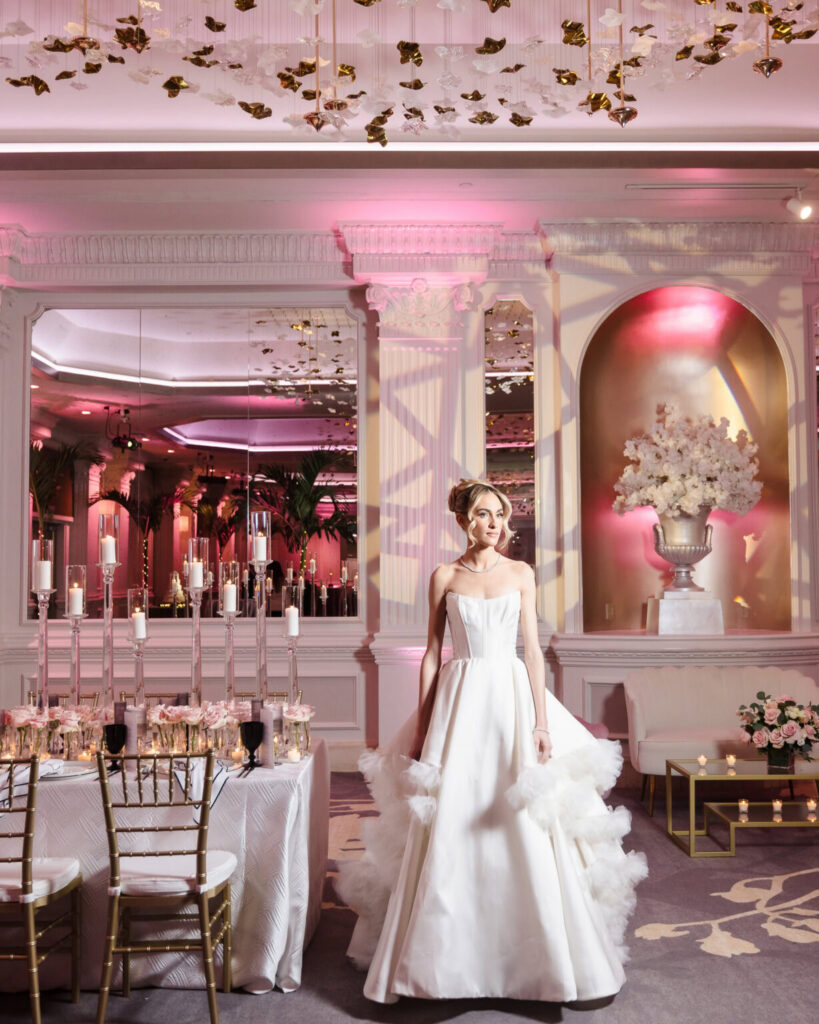 Bride in a modern white wedding gown posing inside an elegant reception ballroom with pink lighting and candlelit tables.