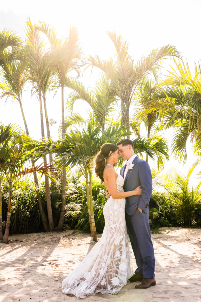 Bride and groom embracing beneath palm trees during a sunlit tropical beach wedding portrait.