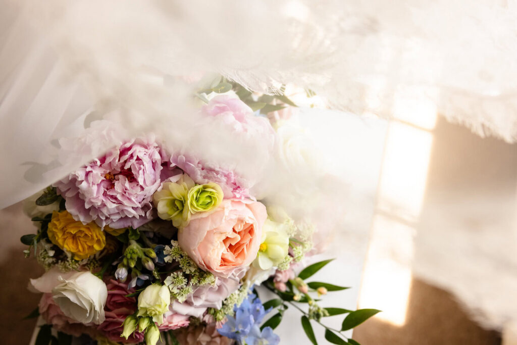 Colorful bridal bouquet with pink, peach, yellow, blue, and white flowers photographed in soft window light.
