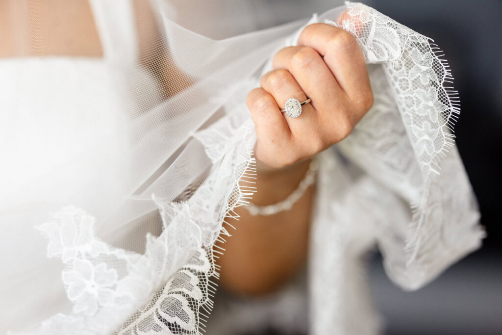 Close-up of a bride’s oval diamond engagement ring framed by the delicate lace edge of her wedding veil showing how much their Long Island wedding cost.