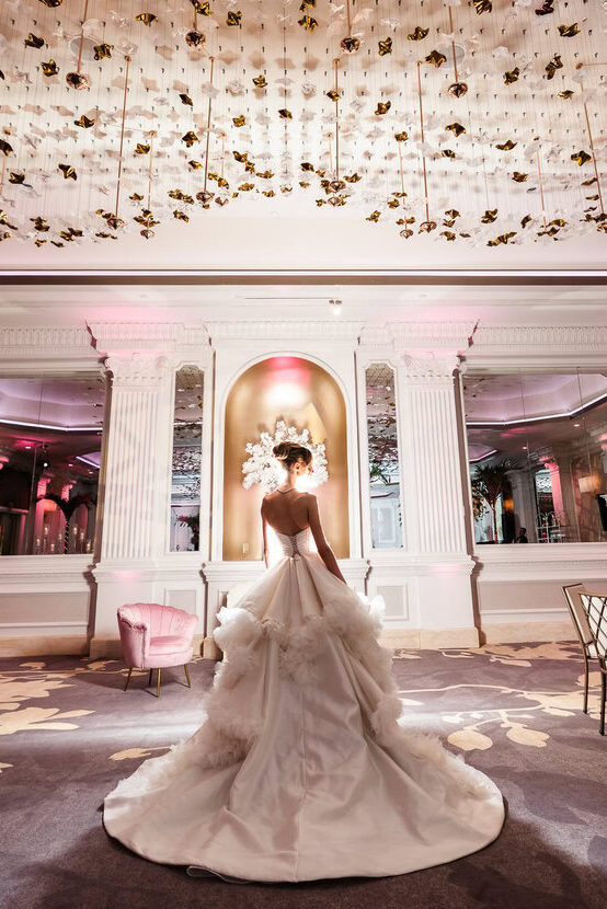 Bride photographed from behind in an elegant ballroom beneath a dramatic ceiling installation and soft pink lighting.