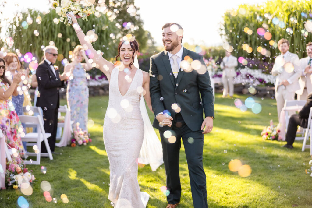Joyful newlyweds walking hand in hand through colorful bubbles after their outdoor wedding ceremony.