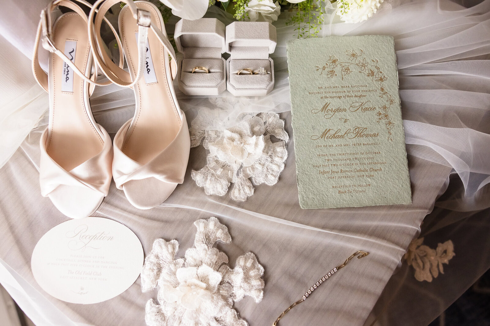 Elegant wedding detail flat lay featuring bridal shoes, wedding rings, invitation suite, lace, and jewelry arranged over a veil.