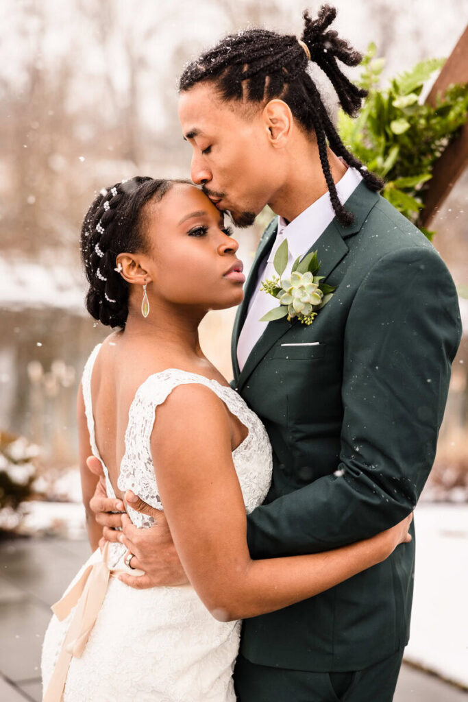 Groom kissing his bride on the forehead as they embrace during a romantic winter wedding portrait in falling snow.