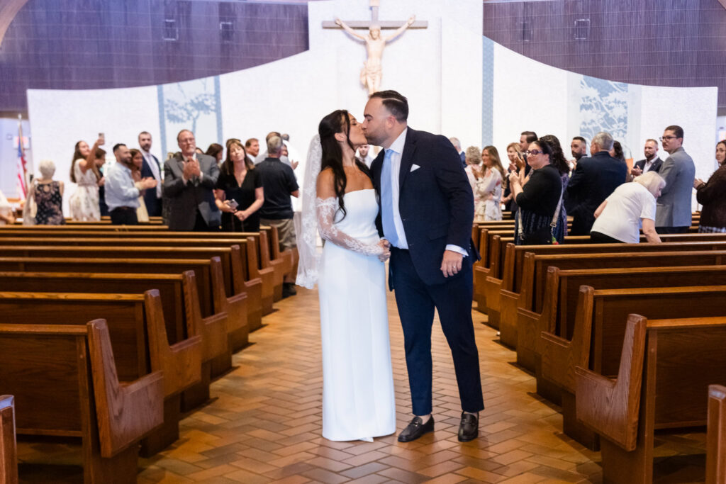 Newlyweds share a kiss in the center aisle of the church as family and friends celebrate behind them.