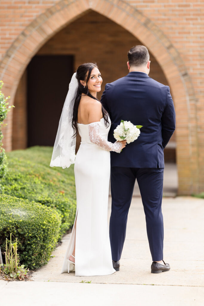 Bride smiles back toward the camera while standing beside the groom beneath a brick archway outside the church.