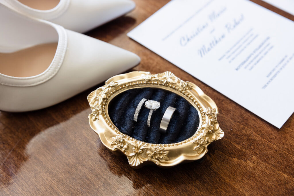 Wedding rings displayed in an ornate gold ring dish beside white bridal shoes and wedding stationery.