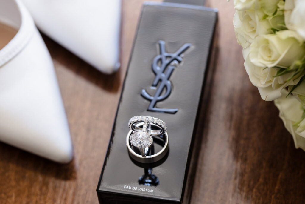 Bride and groom’s wedding rings arranged on a black YSL perfume bottle beside white roses and bridal shoes.