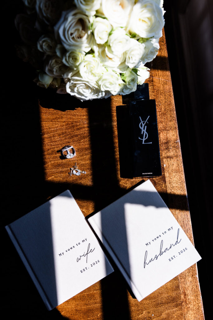 Bride and groom vow books arranged with wedding rings, perfume, jewelry, and a white rose bouquet in window light.
