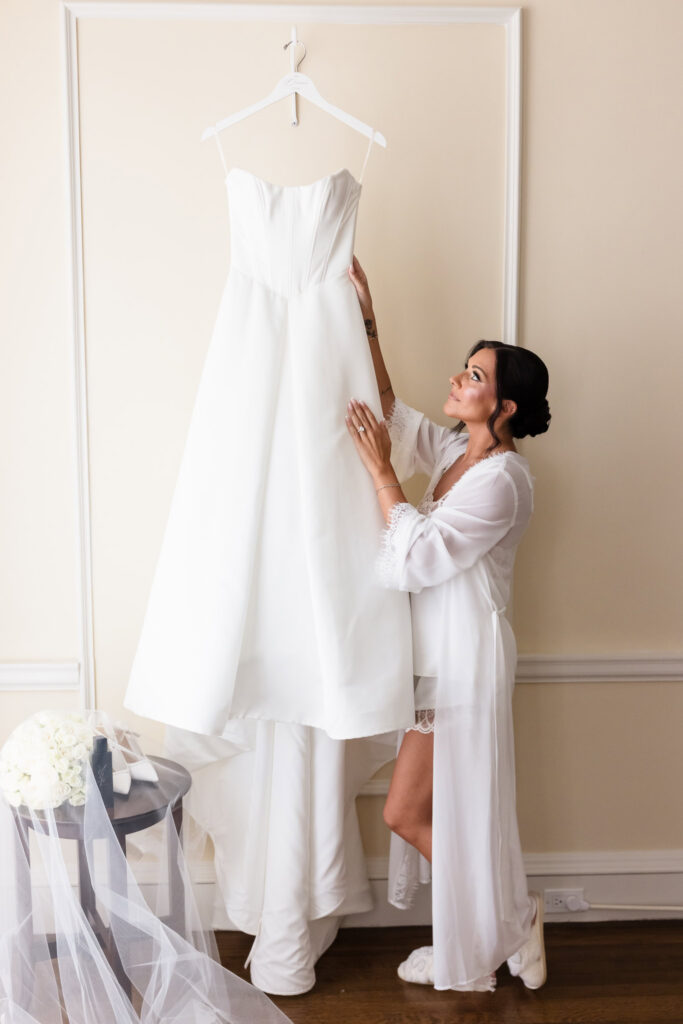 Bride in a white robe looks up at her wedding dress while getting ready before the ceremony.