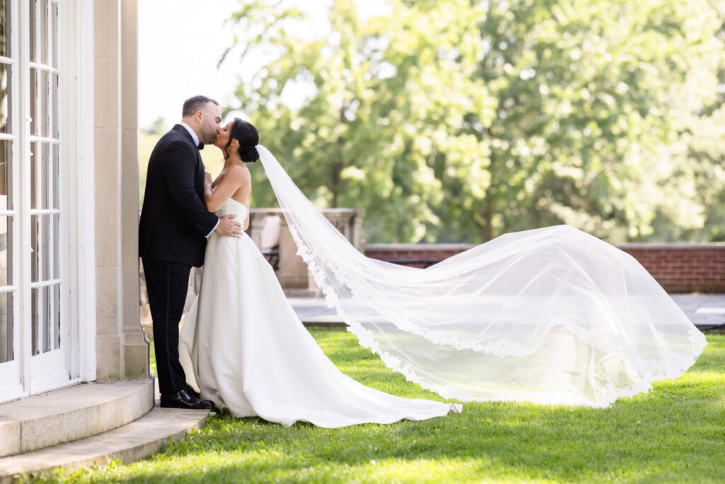 Bride and groom kissing as her long veil flows behind them on the lawn at The Muttontown Club.
