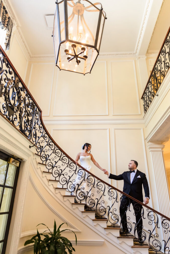 Bride and groom holding hands on the elegant wrought-iron staircase inside The Muttontown Club.