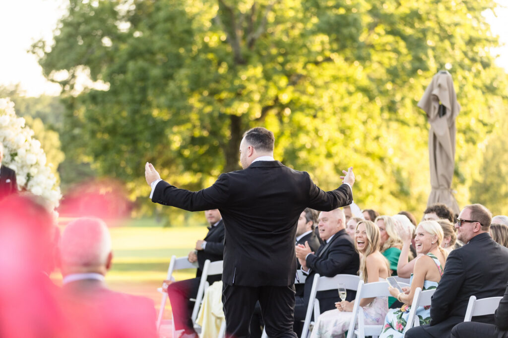 Groom walking down the outdoor ceremony aisle as smiling wedding guests cheer from their seats.