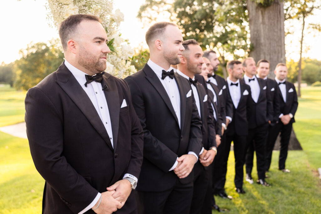 Groom and groomsmen in classic black tuxedos gathered outdoors before the wedding ceremony.