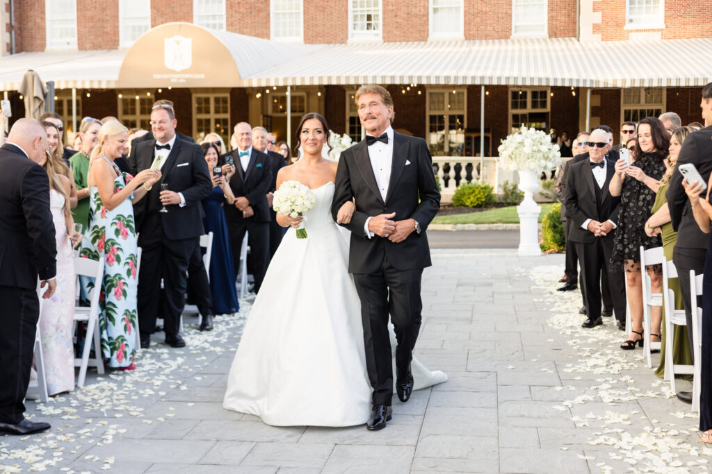 Bride walking down the outdoor wedding aisle with her father surrounded by family and friends.