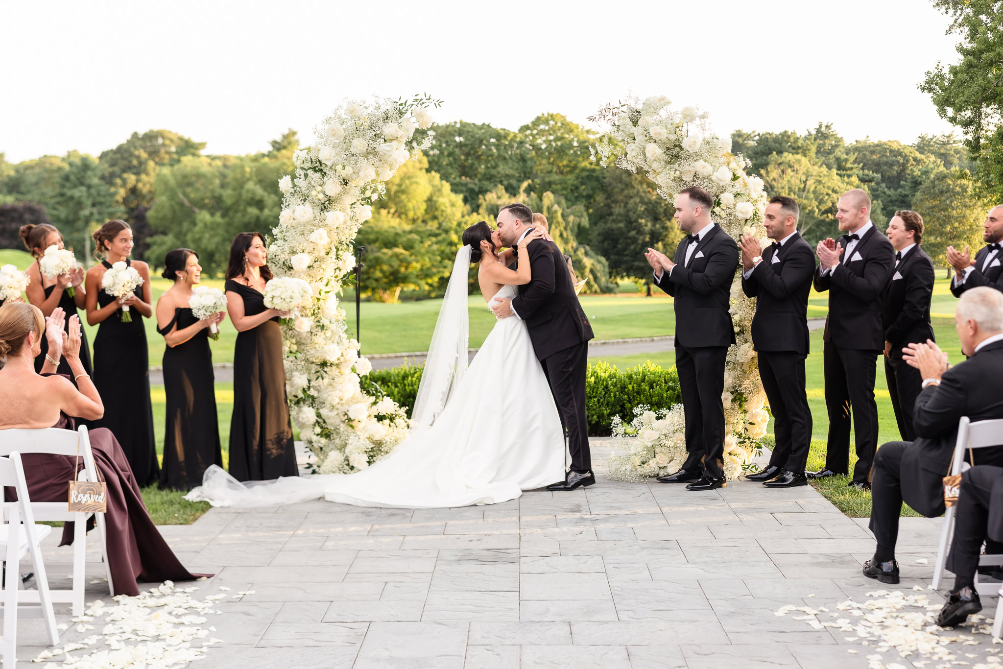 Bride and groom sharing their first kiss beneath a white floral wedding arch during their outdoor ceremony.